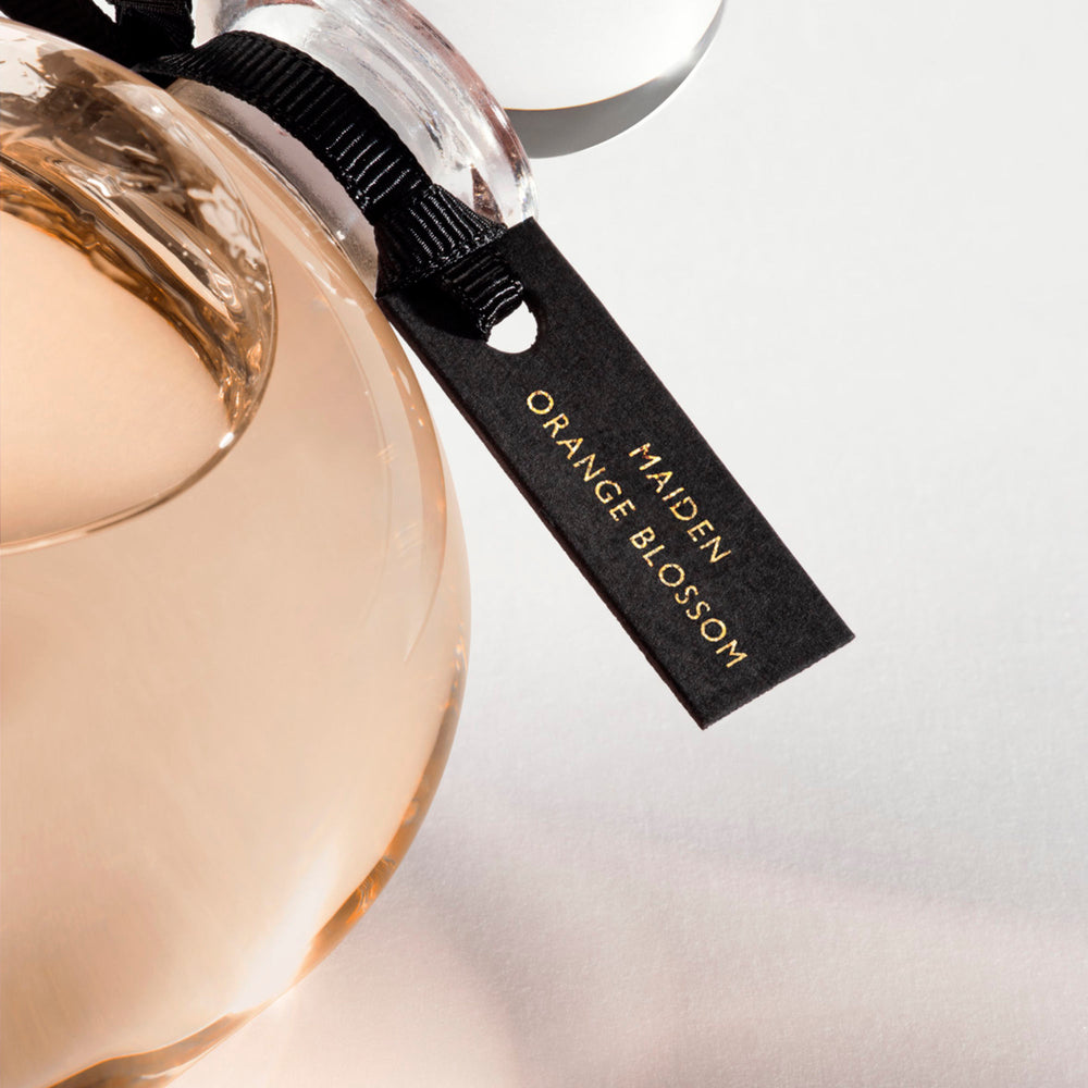 Detail of the top of the 250 ml bottle, with transparent glass and orangey perfum. Spherical cap with black ribbon. Maiden Orange Blossom, a fragrance by Eric Butherbaugh.